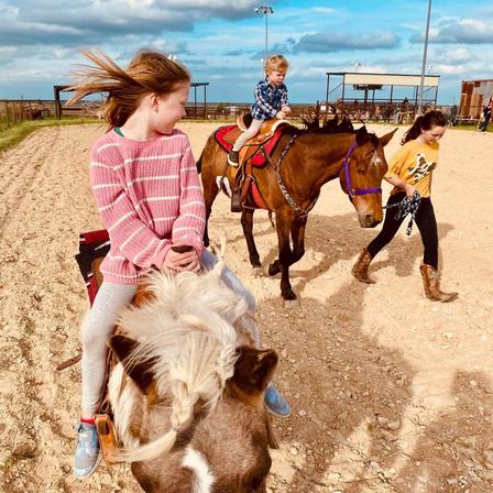 Children ride hand-led ponies around a dirt arena while a handler walks alongside.