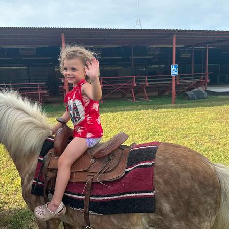 A cheerful toddler in a red top waves from the back of a palomino pony at a festival.