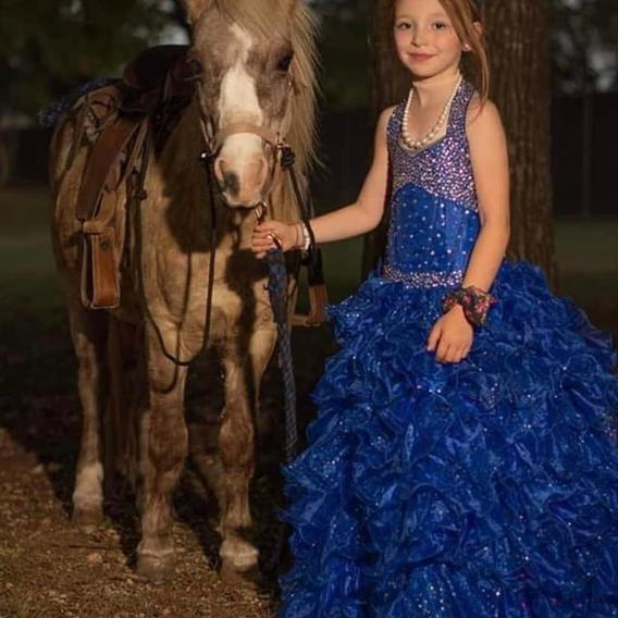 A girl in a sparkling blue ballgown stands with a pony wearing a unicorn horn, photographed on a wooded ranch trail.